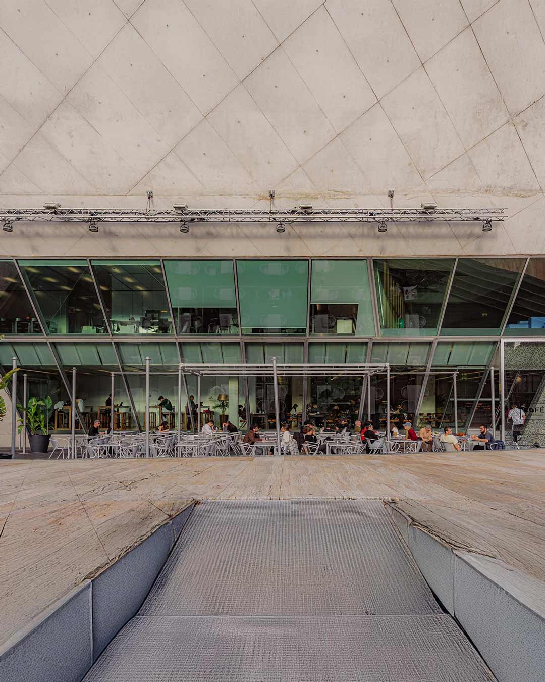 Vista exterior do café MAIS na parte inferior da Casa da Música, durante o dia, com plano amplo que destaca a arquitetura e a esplanada.